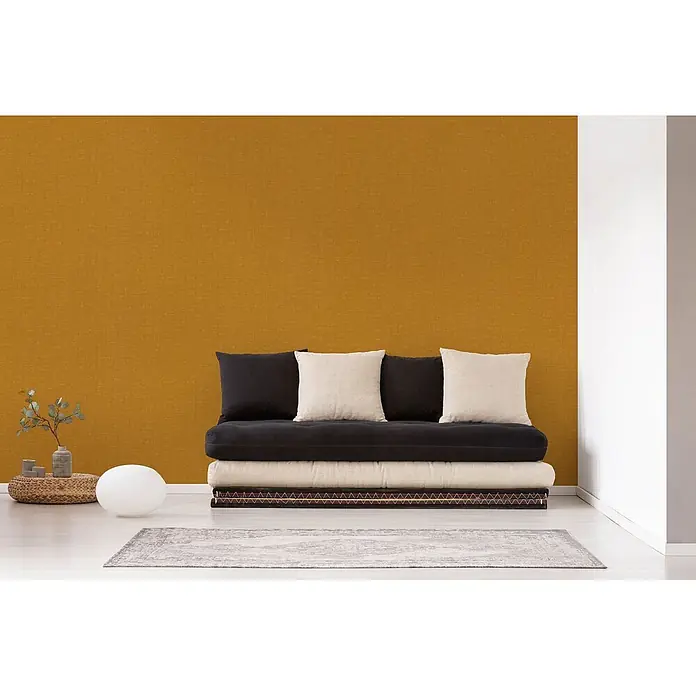 Profhome Vliestapete Heißgeprägte Vliestapete Textiloptik in gelb - ocker-gelb, 10.050 x 530 mm - Schwarzes und beiges Futonsofa mit Kissen in einem Raum mit senfgelber Wand.