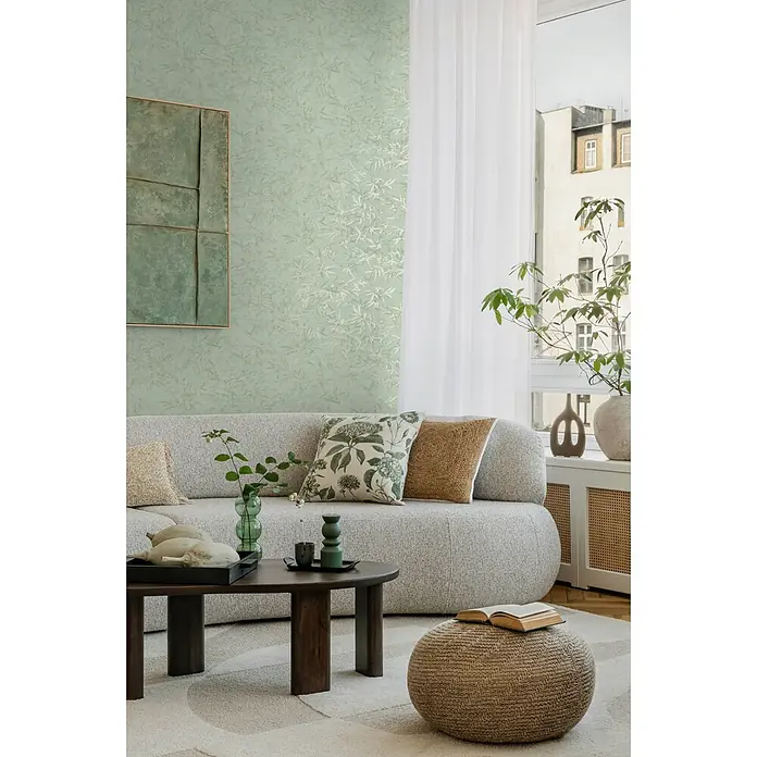 Profhome Vliestapete Heißgeprägte Vliestapete Ton-In-Ton, Floral, Textiloptik in grün - minz-grünsilber, 10.050 x 530 mm - Modernes Wohnzimmer mit hellgrauem Sofa, dunklem Holztisch und geflochtenem Pouf auf gemustertem Teppich, an einer hellgrünen Wand.