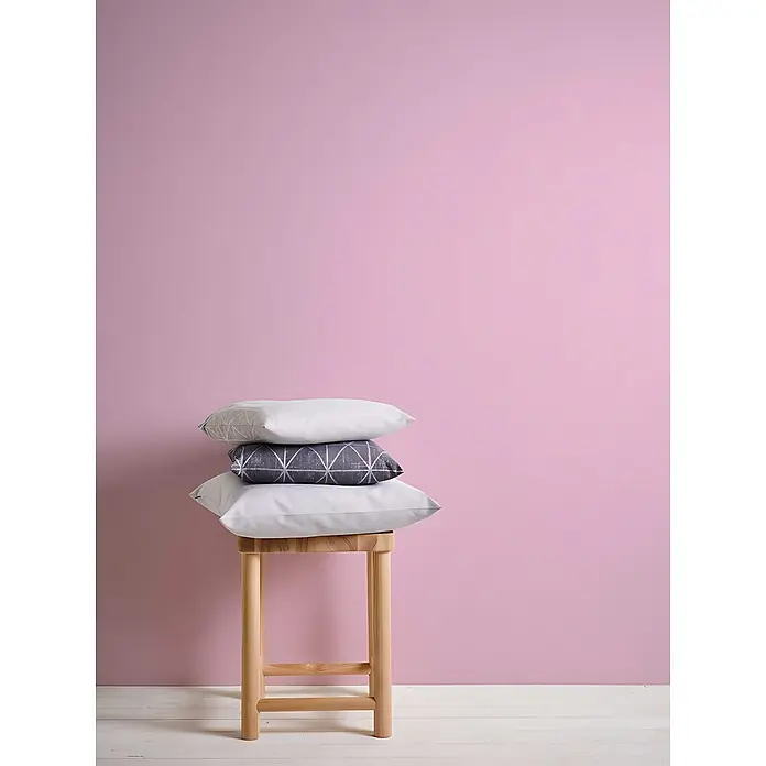 Profhome Vliestapete Vliestapete Uni in rosa - Uni, 10.050 x 530 mm - Drei gestapelte Kissen (zwei weiß, eins grau gemustert) auf einem Holzschemel vor einer rosa Wand.
