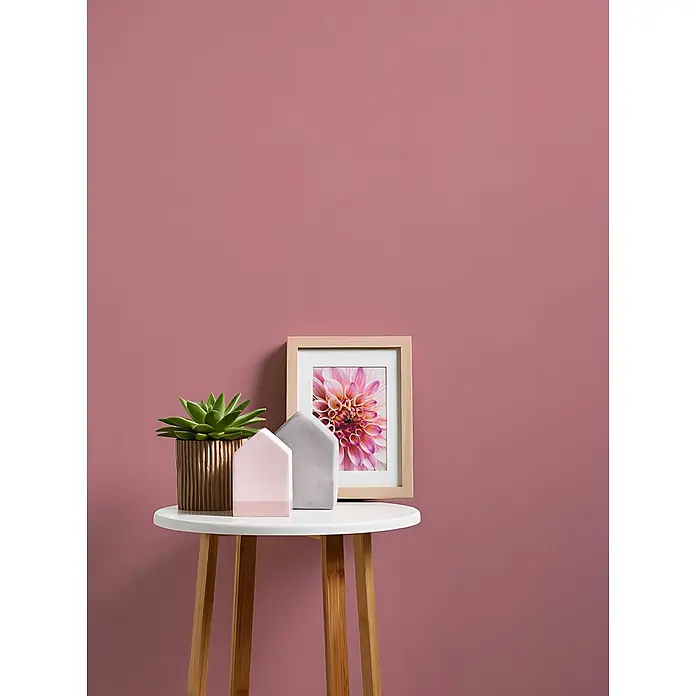 Profhome Vliestapete Vliestapete Uni in rosa - Uni, 10.050 x 530 mm - Kleiner runder Tisch mit grüner Pflanze in bronzefarbenem Topf, rosa und grauen Hausdekorationen und gerahmtem Dahlienbild vor rosa Wand.