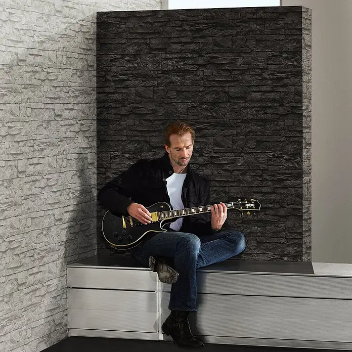 Profhome Vliestapete Vliestapete Steinoptik grau - schwarz, Steinoptik, 10.050 x 530 mm - Mann spielt Gitarre sitzend auf einer silbernen, horizontal geriffelten Aufbewahrungsbank.