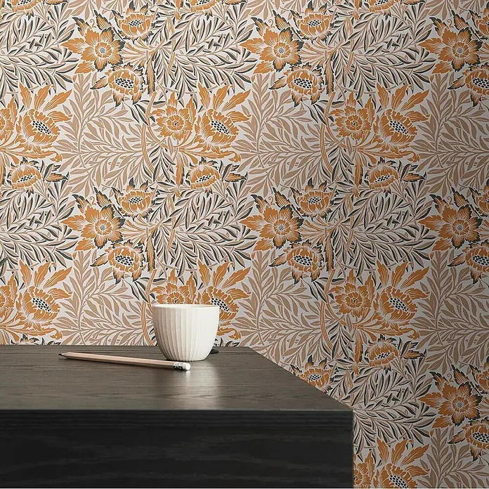 Profhome Vliestapete Vliestapete Blumen, Landhaus, Floral, Retro in orange - beige-braunschwarz, 10.050 x 530 mm - Orange-schwarz gemusterte Blumentapete an einer Wand hinter einem dunklen Holztisch mit Tasse und Bleistift.
