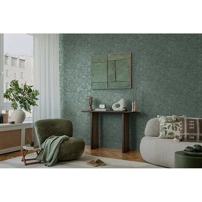 Profhome Vliestapete Heißgeprägte Vliestapete Ton-In-Ton, Floral, Textiloptik in grün - patina-grünplatin, 10.050 x 530 mm - Grüne Tapete mit subtilem Blattmuster, ziert eine Wohnzimmerwand hinter Möbeln.
