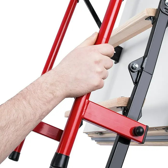 Bodentreppe Polar Plus - 120 x 70 cm, Wärmedämmung: 0,36 W/(m²K) - Eine Hand hält die rote Metallleiter einer Dachbodentreppe. Die Treppe ist teilweise ausgeklappt und an einer weißen Fläche befestigt.