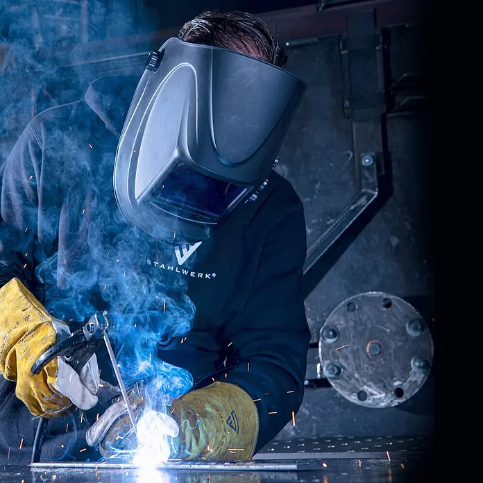 STAHLWERK Kombi-Schweißgerät ARC 200 Synergic Pro MMA Lift TIG - Schweißstrom: 200 A - Schweißer mit schwarzem Helm, Hoodie und gelben Handschuhen beim Schweißen mit hellen Funken und Rauch.