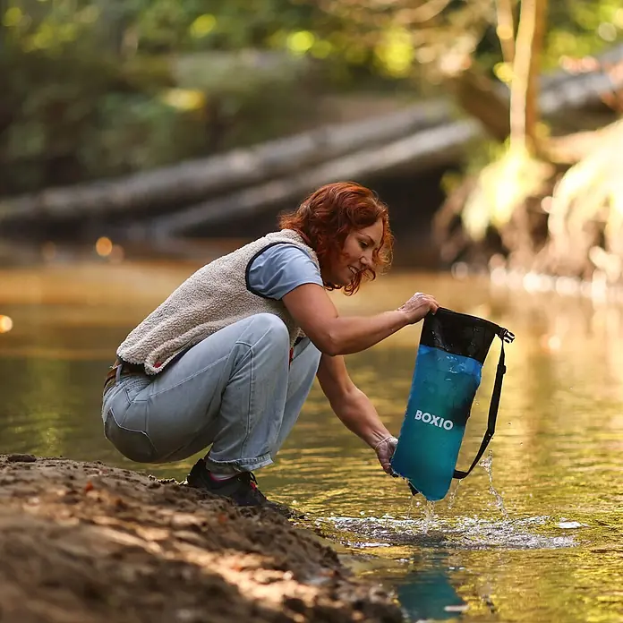 Boxio Campingwaschbecken Wash Max+ - 40 x 30 x 40 cm, 5 -tlg. - Frau in der Natur füllt einen blau-schwarzen BOXIO Packsack mit Wasser aus einem Fluss.