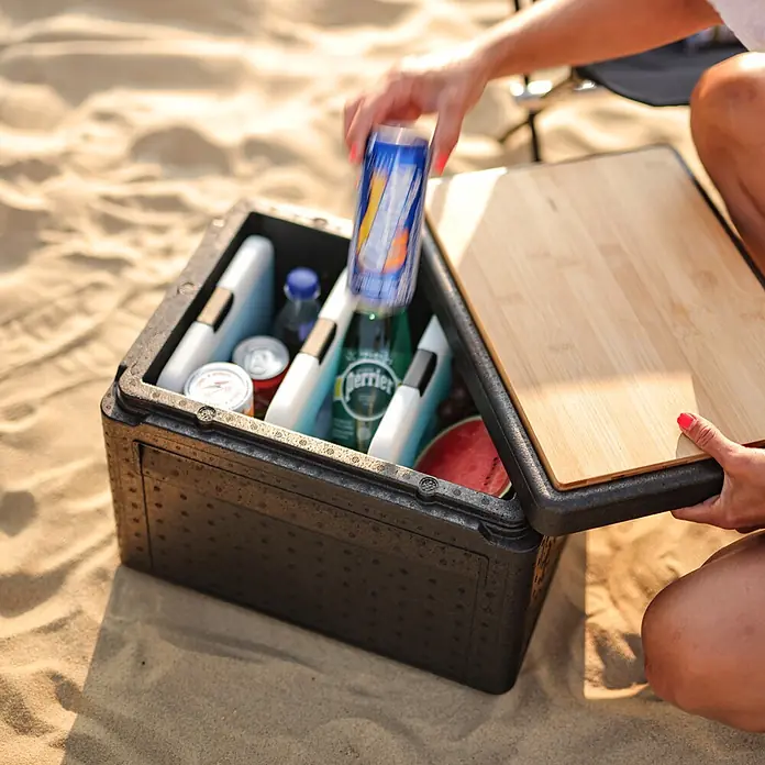 Boxio Kühlbox Cool - Schwarz, 16 l, 1 -tlg. - Schwarze Kühlbox mit Holzdeckel, gefüllt mit Getränken und Snacks, am Sandstrand. Eine Hand legt eine Dose hinein.