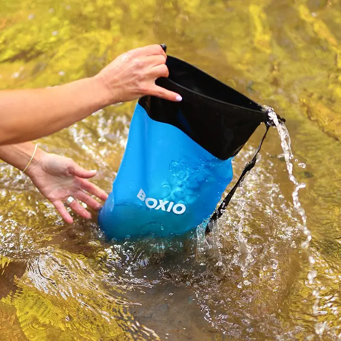 Boxio Wasserfilter Set - 5 -tlg. - Hände tauchen eine blau-schwarze BOXIO-Trockentasche in spritzendes Wasser und zeigen ihre Wasserdichtigkeit.