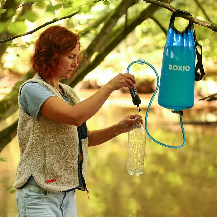 Boxio Wasserfilter Set - 5 -tlg. - Eine Frau füllt eine Flasche Wasser aus einem blauen tragbaren Filtersystem, das in einem Wald hängt.
