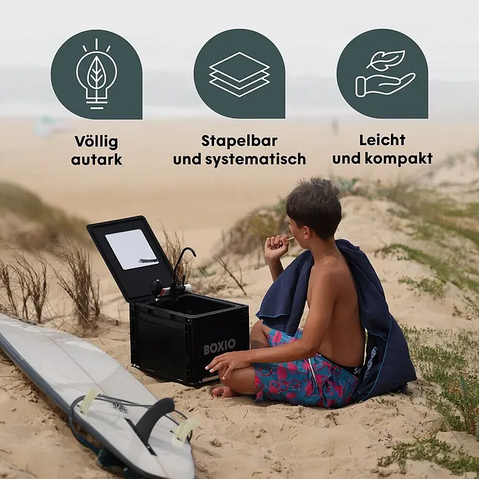 Boxio Campingwaschbecken Wash - 40 x 30 x 28 cm, 1 -tlg. - Schwarze, tragbare Outdoor-Waschstation 'BOXIO' mit Wasserhahn, von einem Jungen am Strand neben einem Surfbrett benutzt.