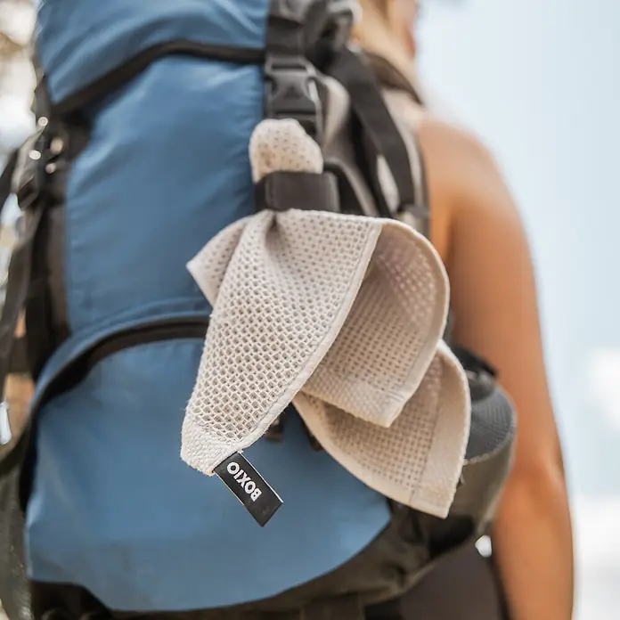 Boxio Campingwaschbecken Wash Max+ - 40 x 30 x 40 cm, 5 -tlg. - Hellbeiges Waffelstrick-BOXIO-Handtuch an einem blauen Rucksackriemen, von einer Person im Freien getragen.