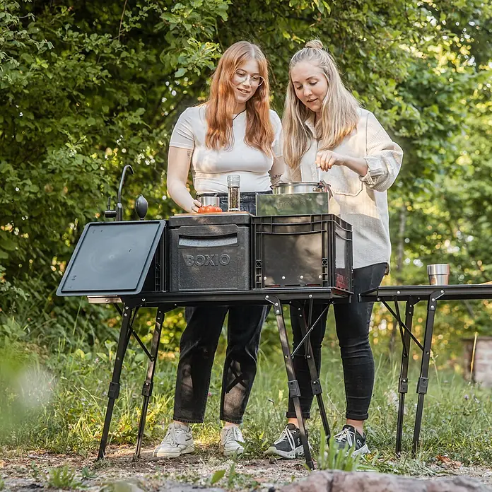 Boxio Campingtisch - L x B x H: 90 x 21 x 11,5 cm, Aluminium - Zwei Frauen kochen draußen an einer schwarzen, tragbaren Outdoorküche mit integrierten Kisten und Spüle.