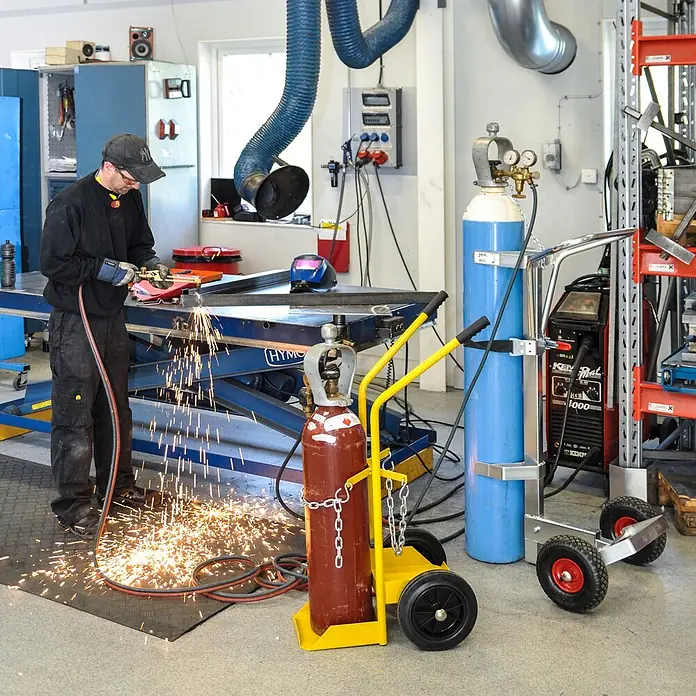 Kongamek Sackkarre Gasflaschenwagen 530x1080x550mm für 50l-Fässer mit 80kg Tragkraft - Traglast: 150 kg - Mann trennt Metall mit einem Brennschneider, Funken fliegen. Gasflaschen auf gelben und silbernen Rollwagen stehen bereit.