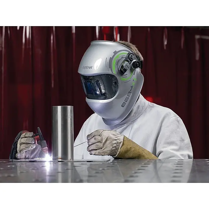 Optrel Schweißhelm E684 -  - Eine Person trägt einen silbernen Optrel Schweißhelm mit grünen Akzenten und schweißt einen Metallzylinder auf einer Werkbank.