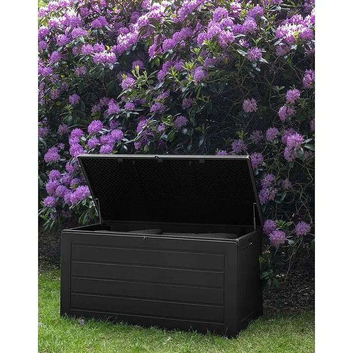 Ebuy24 Garten-Aufbewahrungsbox Dubrovnik - L x B x H: 61 x 147 x 65 cm, Schwarz, Kunststoff - Dunkle Anthrazit-Gartenbox mit geöffnetem Deckel, auf Rasen vor blühendem Strauch.