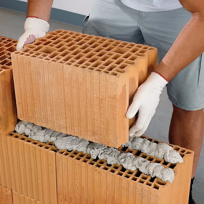 IsolBau Klebeschaum PU Mauermörtel - 6 x 750ml, PU-Reiniger - 500 ml, Schaumpistole - 750 ml - Arbeiter setzt großen, hohlen terrakottafarbenen Keramikziegel mit grauem Klebemörtel.