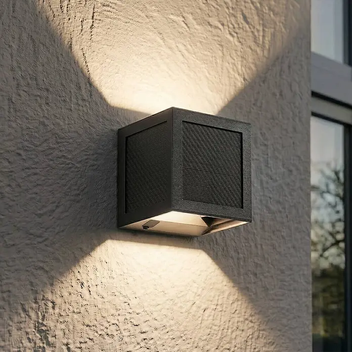 Prios Außenwandleuchte Avvin - 10 x 10 x 10 cm, schwarz, schwarz, IP65 - Dunkelgraue quadratische Außenwandleuchte mit strukturierter Umrandung und Gitterdetails, wirft Licht nach oben und unten an eine strukturierte Wand.
