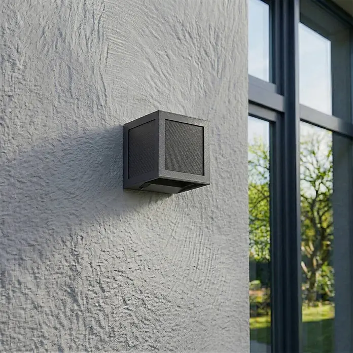 Prios Außenwandleuchte Avvin - 10 x 10 x 10 cm, schwarz, schwarz, IP65 - Dunkelgraue, quadratische Außenwandleuchte an einer strukturierten Außenwand neben einem Fenster.