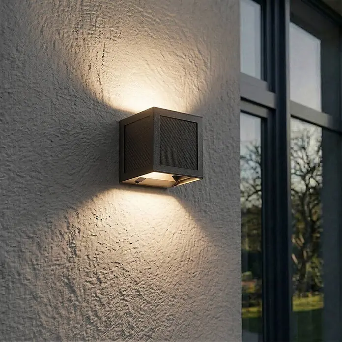 Prios Außenwandleuchte Avvin - 10 x 10 x 10 cm, schwarz, schwarz, IP65 - Dunkelgraue, quaderförmige Außenwandleuchte an einer strukturierten Wand, die Licht nach oben und unten wirft, neben einem Fenster.