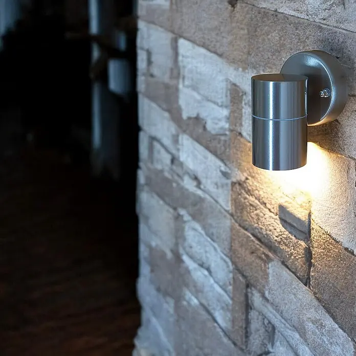 DAPO Außenwandleuchte Bornholm, Silber, Edelstahl, GU10, IP44 -  - Zylindrische Wandleuchte aus gebürstetem Metall, an einer Steinwand montiert, strahlt warmes Licht nach unten ab.