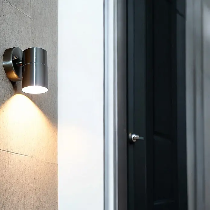 DAPO Außenwandleuchte Bornholm, Silber, Edelstahl, GU10, IP44 -  - Zylindrische Wandleuchte aus gebürstetem Metall, strahlt warmes Licht nach unten auf eine strukturierte Wand neben einer dunklen Tür.