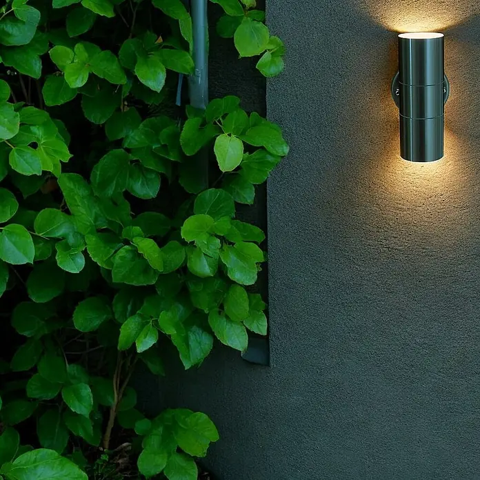 DAPO Außenwandleuchte Bornholm Up&Down, Edelstahl, 2-Flammig, GU10, IP44 -  - Gebürstete Edelstahl-Wandleuchte mit Auf-/Ablichtung, die warmes Licht auf eine dunkle Wand neben grünen Pflanzen wirft.