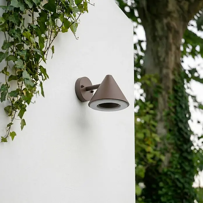Lindby Außenwandleuchte Soran - braun / rost, braun / rost, IP44 - Braune konische Außenwandleuchte an weißer Wand, mit grünem Efeu und verschwommenem Baum im Hintergrund.