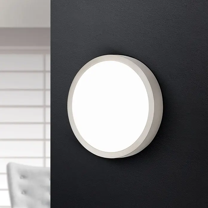 Orion Leuchten LED-Panel rund -  - Runde LED-Wandleuchte mit silbernem Rahmen und beleuchteter weißer Fläche, an einer dunklen Wand montiert.