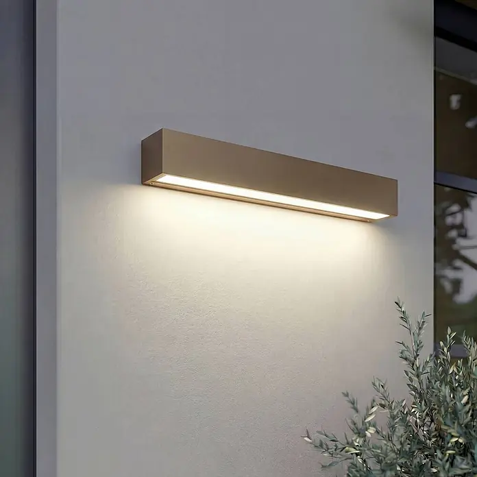 Lucande Außenwandleuchte Lengo - 6 x 50 x 6 cm, braun / rost, IP65 - Rechteckige bronzefarbene Außenwandleuchte beleuchtet eine Wand mit warmem Licht.