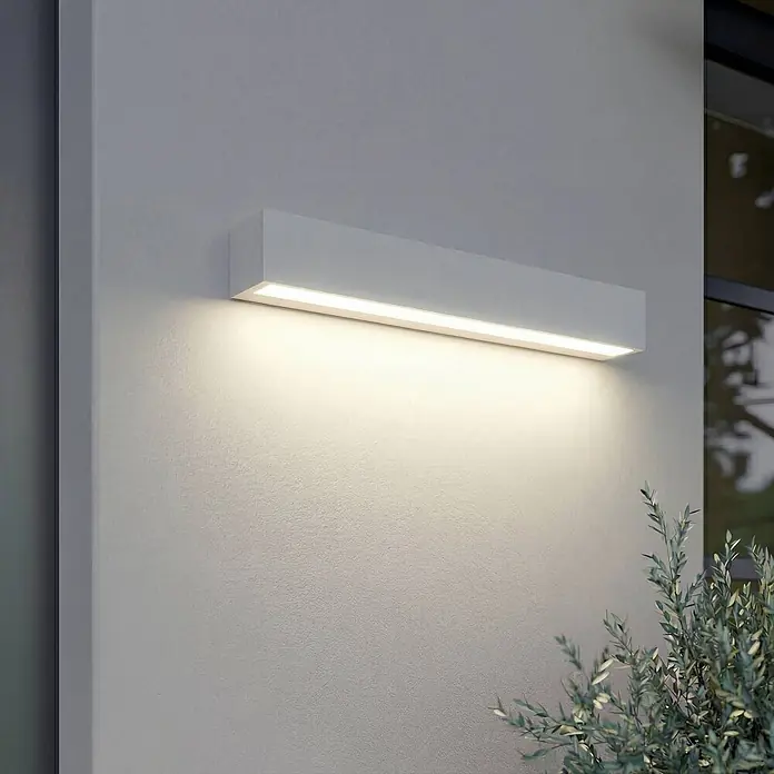 Lucande LED Wandleuchte aussen in weiß, Aussenlampe Wand -  - Weißes, rechteckiges Außenwandlicht leuchtet mit warmem Licht nach unten auf eine strukturierte Wand neben einer grünen Pflanze.