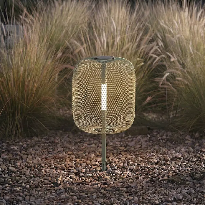 Lucande LED Solar Gartenlampe Erdspieß in grün, dimmbar Erdspießlampe -  - Eine beleuchtete, hellfarbene Mesh-Gartenleuchte mit Solarpanel, in Kies zwischen hohem Gras gesteckt.