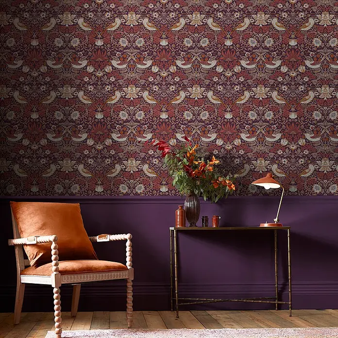 William Morris At Home Vliestapete Erdbeerdieb - Lila, Floral, 1.000 x 52 cm - Dunkelviolette Tapete mit sich wiederholenden Vogel- und Blumenmustern in Rost, Beige und Orange, die eine Zimmerwand bedeckt.