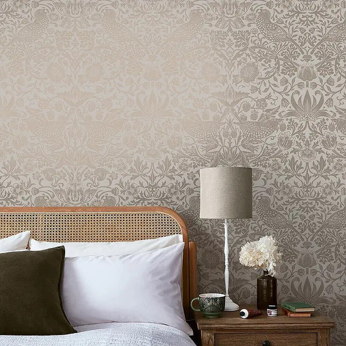 William Morris At Home Vliestapete Erdbeerdieb - Beige, Floral, 1.000 x 52 cm - Beige/taupe Tapete mit aufwendigem Blumen- und Vogelmuster an einer Schlafzimmerwand über einem Rattan-Kopfteil.