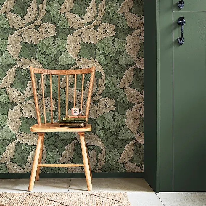 William Morris At Home Vliestapete Vliestapete Akanthus - Grün, Streifen, 10 x 0,52 m - Grüne und beige Tapete mit botanischem Blattmuster an der Wand hinter einem Holzstuhl.