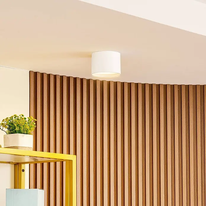 Lindby Deckenstrahler Nivoria - weiß / opal - Weiße zylindrische Deckenleuchte an einer cremefarbenen Decke über einer geschwungenen Holzlamellenwand.