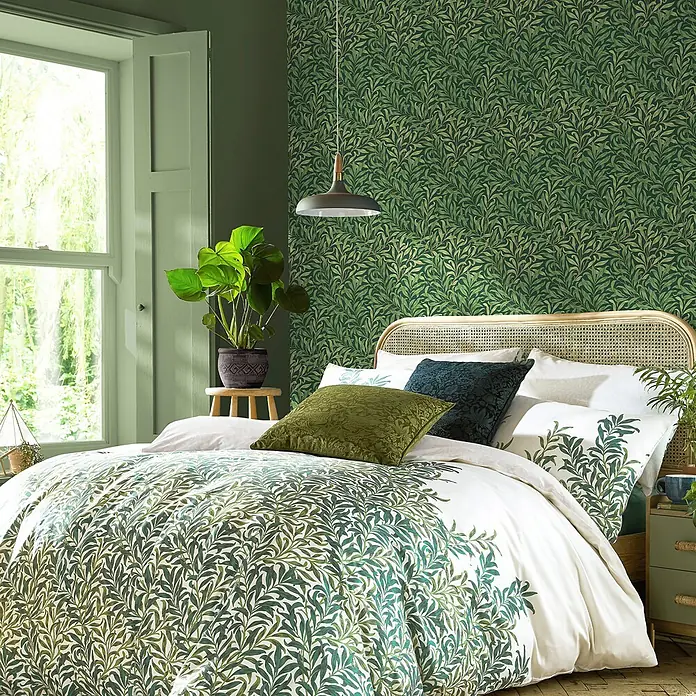 William Morris At Home Vliestapete Weidenzweig - Grün, Floral, 1.000 x 52 cm - Bett mit weißer Bettwäsche und Kissenbezügen mit üppigem grünem Blattmuster.