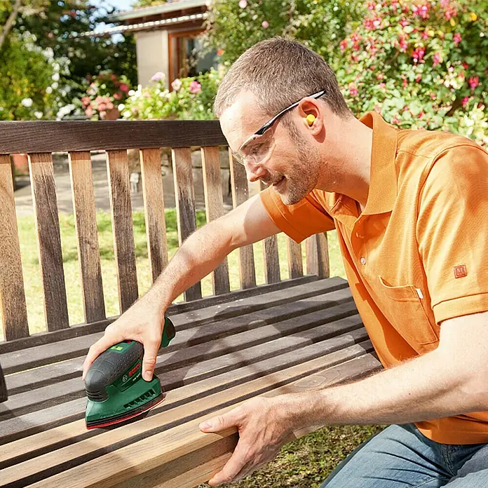Bosch Lijadora delta de batería EasySander 12 - 12 V, Iones de litio, Sin batería, 22.000 r.p.m. - Un hombre lija un banco de madera al aire libre con una lijadora eléctrica Bosch. Lleva gafas de seguridad y tapones para los oídos.