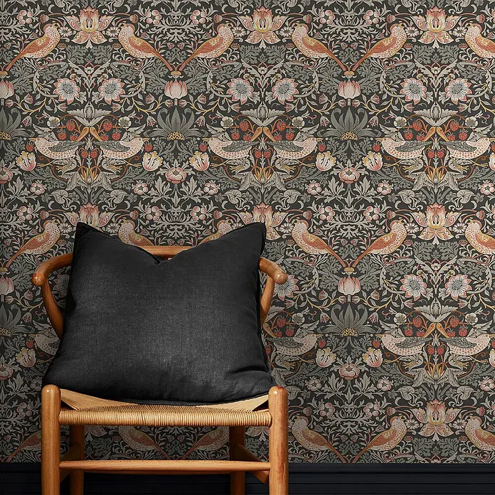 William Morris At Home Vliestapete Erdbeerdieb - Grau, Floral, 1.000 x 52 cm - Schwarzes Stoffkissen auf einem Holzstuhl vor einer dunklen Tapete mit Blumen- und Vogelmuster.
