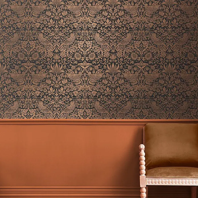 William Morris At Home Vliestapete Vliestapete Erdbeerdieb - Schwarz/Kupfer, Floral, 10 x 0,52 m - Dunkle Tapete mit wiederholtem kupferfarbenem Blumen- und Vogelmuster, über orangefarbener Vertäfelung, mit einem Stuhl sichtbar.