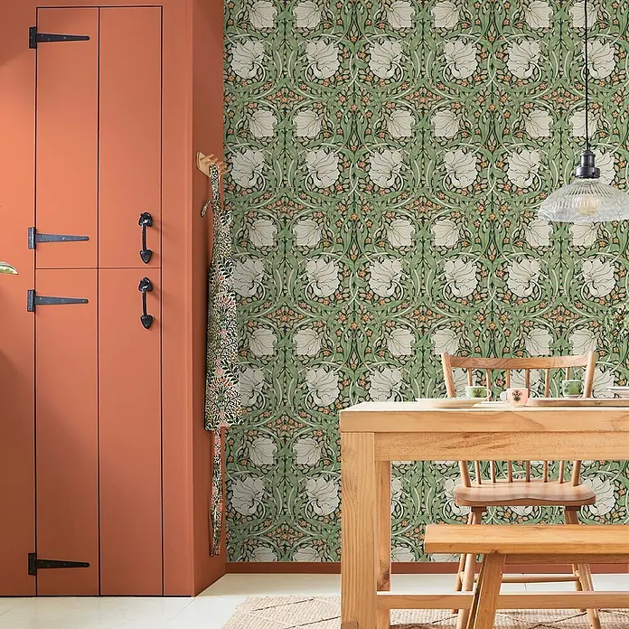 William Morris At Home Vliestapete Vliestapete Pimpernel Blumen - Grün, Floral, 10 x 0,52 m - Grüne Blumentapete in einem Raum mit einem orangebraunen Schrank und einer hellen Holz-Essgruppe.