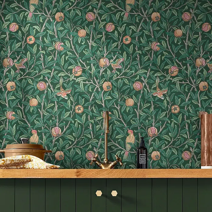 William Morris At Home Vliestapete Vliestapete Vogel und Granatapfel - Grün, Tieroptik - Dunkelgrüne Tapete mit lebhaftem Muster aus Vögeln, Granatäpfeln und Blättern an einer Küchenwand über einer Holztheke.