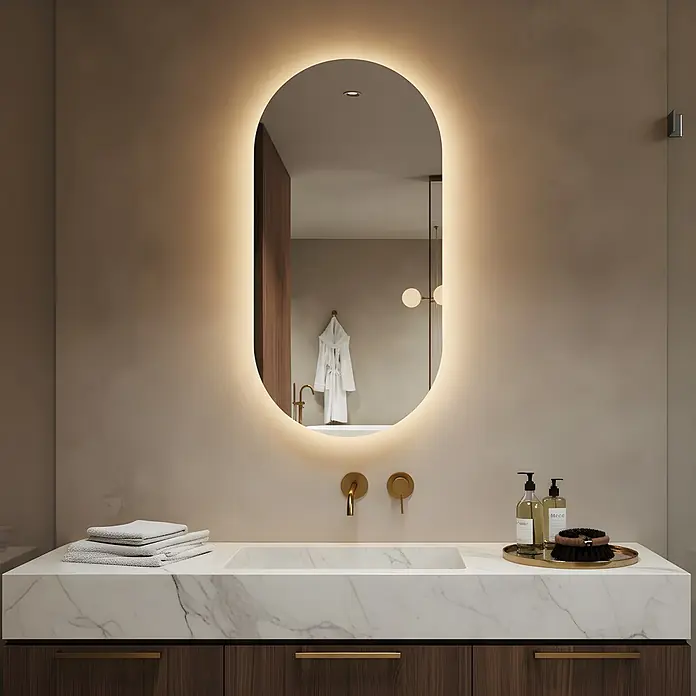 Lumoire Lichtspiegel LUX - 50 x 90 cm - Modernes Badezimmer mit ovalem, beleuchtetem Spiegel über einem Waschtisch aus weißem Marmor, mit Messingarmaturen und dunklen Holzschränken.