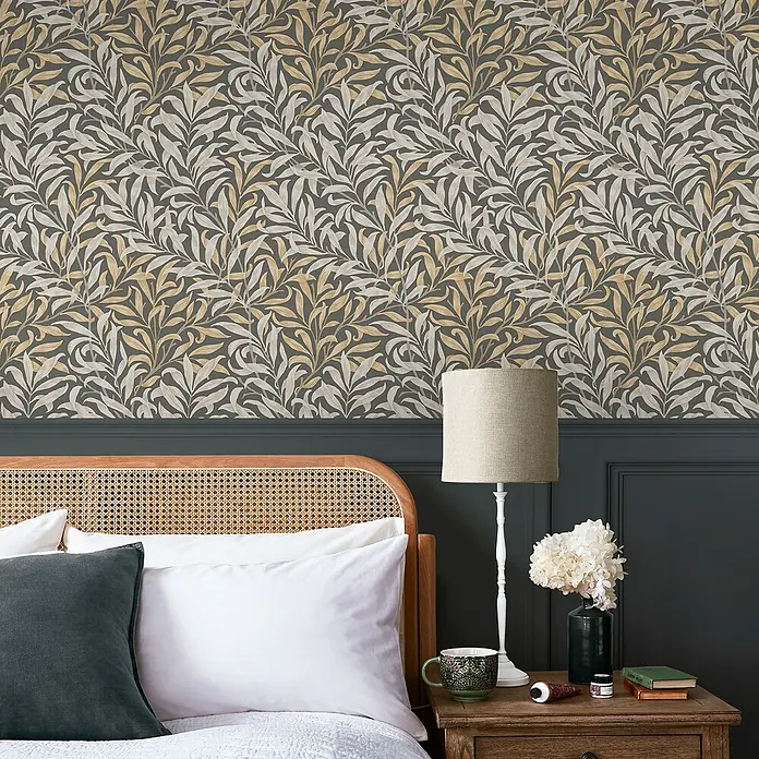 William Morris At Home Vliestapete Weidenzweig - Grau, Floral, 1.000 x 52 cm - Tapete mit dunkelgrauem Hintergrund und elegantem Muster aus hellbeigen und mattgoldenen Blättern an einer Schlafzimmerwand.
