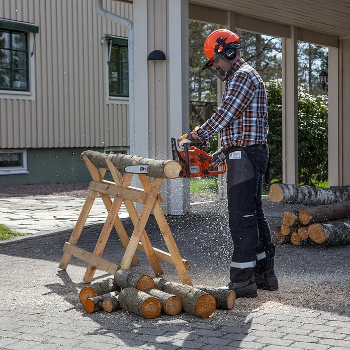 Husqvarna Benzin-Kettensäge 120 Mark II - 1.400 W, 38 cm³, Schwertlänge: 35 cm, Max. Aststärke: 300 mm - Mann in Schutzkleidung sägt mit Motorsäge ein Holzscheit auf einem Sägebock, Späne fliegen.