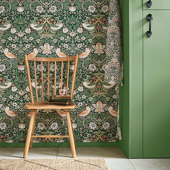 William Morris At Home Vliestapete Vliestapete Erdbeerdieb - Dunkelgrün, Floral, 10 x 0,52 m - Dunkelgrüne Tapete und passender Stoff mit detailliertem Vogel-, Erdbeer- und Blumenmuster, neben einem Holzstuhl.