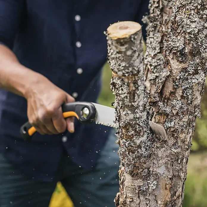 Fiskars Plus Klappsäge SW69 - 21 cm - Person sägt mit einer Handsäge mit silberner Klinge und orange/schwarzem Griff einen Ast ab.