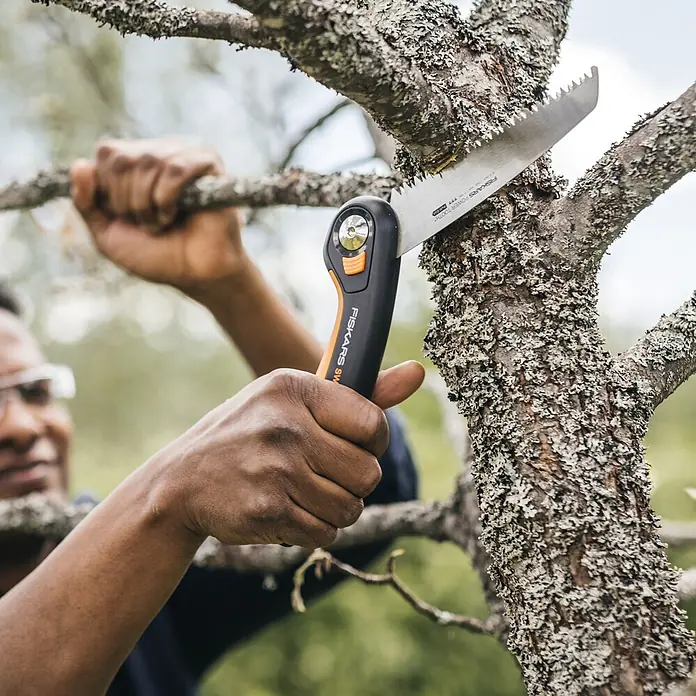 Fiskars Plus Klappsäge SW68 - 15 cm - Eine Person nutzt eine Fiskars Astsäge mit orange-schwarzem Griff, um einen lichenbedeckten Ast zu sägen.