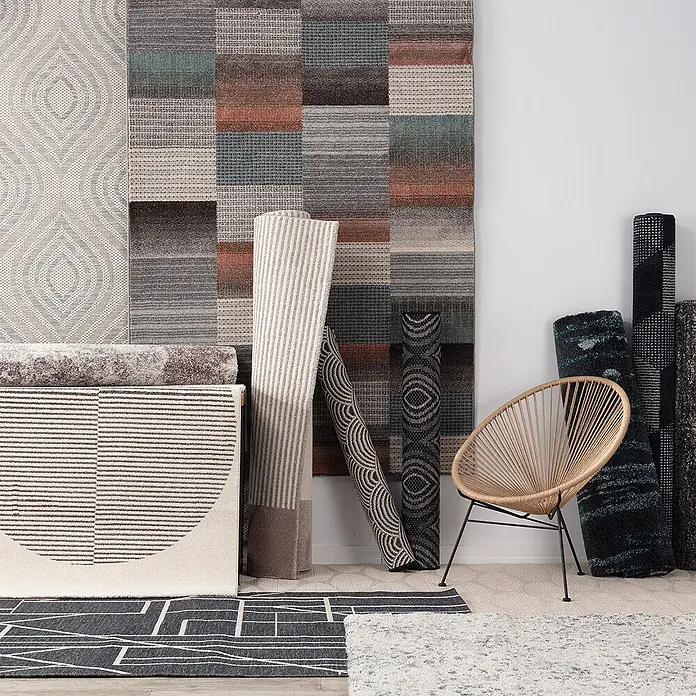 Dekoria Teppich Breeze beige/raw/blue 200x290cm -  - Eine Auswahl an gemusterten und texturierten Teppichen, teils gerollt, teils ausgelegt, präsentiert mit einem modernen Korbstuhl.