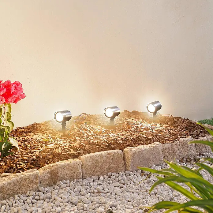 Lindby Erdspießleuchte Nycta -  - Drei silberne Außenstrahler beleuchten ein Blumenbeet mit warmem Licht.
