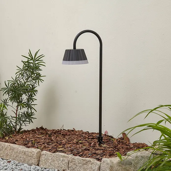 Lindby Erdspießleuchte Ameline - 77,2 cm - Eine dunkelgraue Wegeleuchte für den Außenbereich mit gebogenem Stiel und geripptem konischem Schirm, die ein Gartenbeet mit Rindenmulch und Pflanzen beleuchtet.